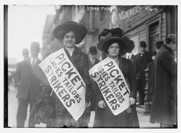 Two women wearing large hats and sashes that read Picket Ladies Tailors Strikers stand on a city street.