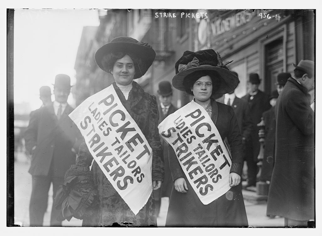 Two women wearing large hats and sashes that read Picket Ladies Tailors Strikers stand on a city street.